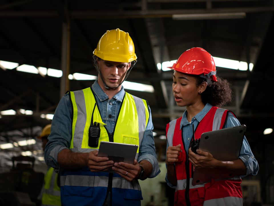 Dois trabalhadores de capacete e colete refletivo conferem um tablet no galpão
